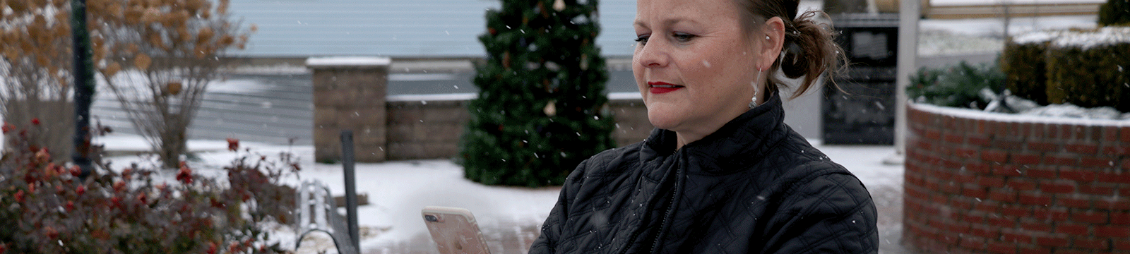 Woman looking at her digital banking on a smartphone outside in the snow. A decorated Christmas tree is in the background.