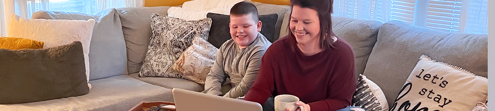 Woman holding a coffee mug, sitting on her couch with son, viewing her laptop.