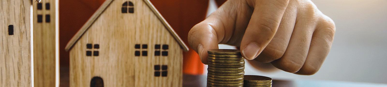 Close up of hand stacking pennies with wooden house models in the background.
