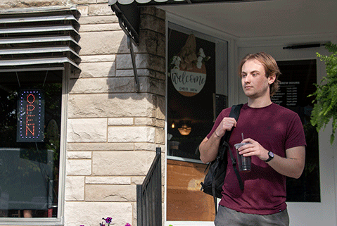 Student wearing a backpack and exiting local coffee shop with an iced coffee.