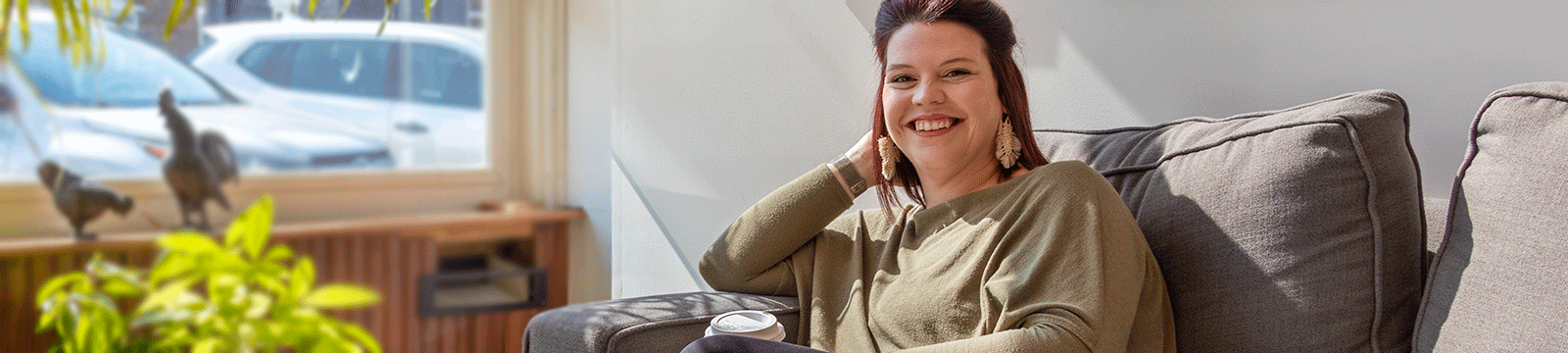 Woman smiling, sitting on a couch, and drinking coffee.